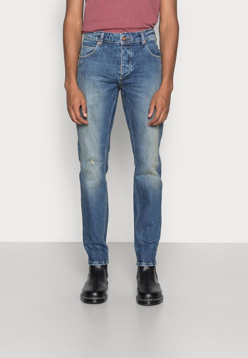 Vaqueros de mezclilla azul con corte recto, parches desgastados, ligeras marcas de desgaste, cinco bolsillos y costuras en contraste, combinados con botas negras hasta el tobillo.