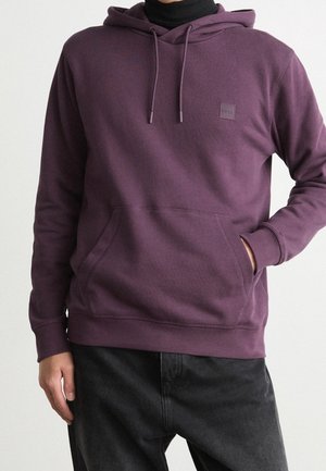 Persoon die een pruimkleurige hoodie draagt over een zwarte coltrui, met één hand in de buidelzak aan de voorkant, gecombineerd met een zwarte broek.