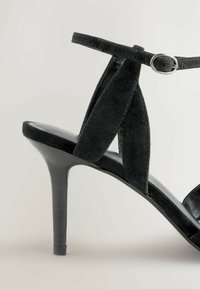 Sort ruskinds højhælet sandal med en slank stiletto hæl, åben tå-design og ankelrem med metalspænde.