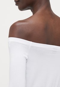 Hvit off-the-shoulder topp laget av myk stoff, med lange ermer og en jevn tekstur, som fremhever skuldre og krageområde.
