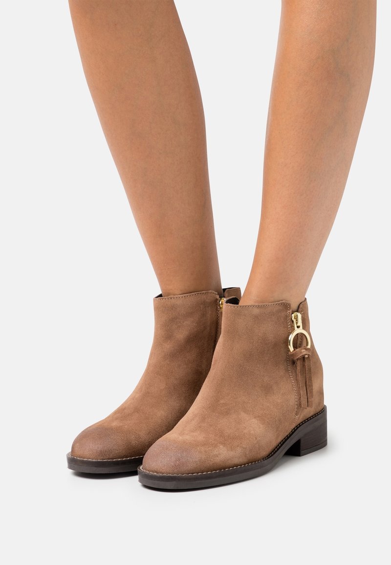 Bottines en daim brun à talons bas, avec fermetures éclair latérales et tirettes de fermeture en forme d'anneaux dorés, portées par une personne aux jambes nues.
