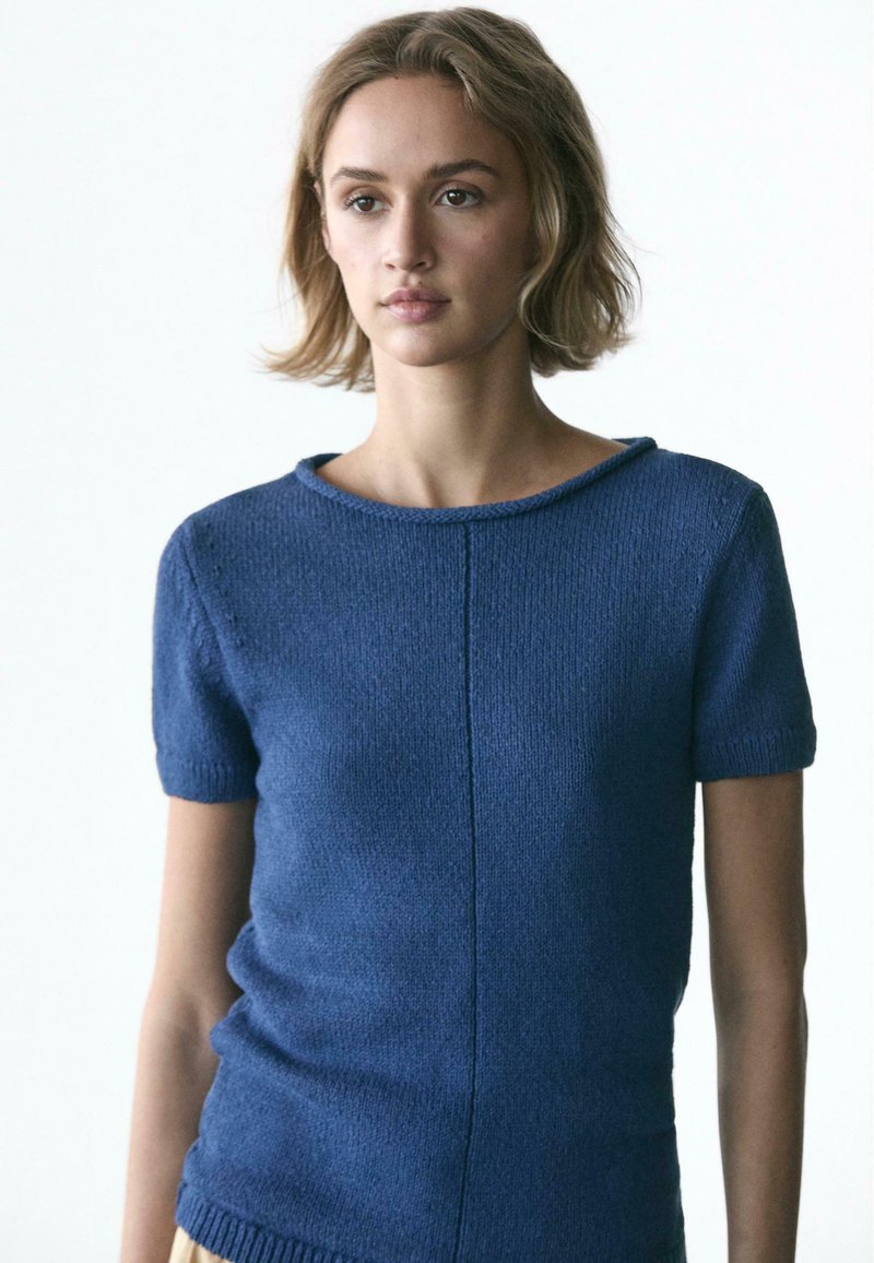 Jeune femme aux cheveux blonds courts, portant un haut en maille bleu à manches courtes, posant devant un fond clair uni.