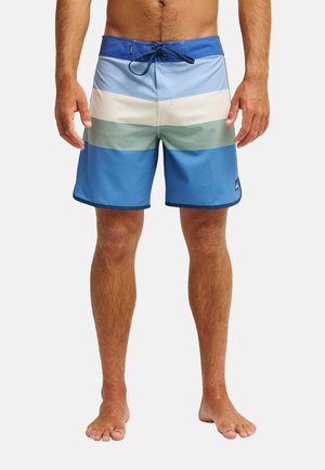 Mann mit blau-weiß-grün gestreiften Badeshorts mit Kordelzug, barfuß stehend auf einem schlichten weißen Hintergrund.