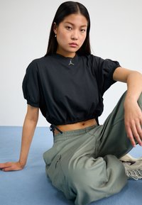 Black cropped top with puff sleeves and drawstring hem, paired with loose green cargo pants. Features a small logo on the neckline.