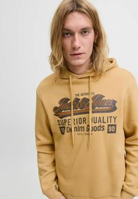 Mustár sárga pulóver kapucnival, elülső grafikával, amelyen a "Jack & Jones" logó és sötétbarna szöveg látható; állítható zsinór a kapucnin.