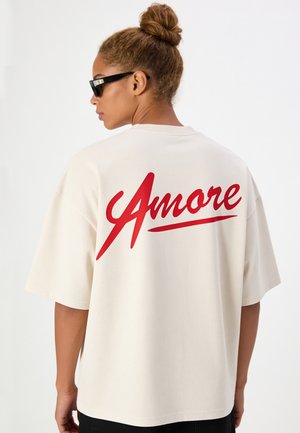 Vrouw met zonnebril en een knot als kapsel draagt een wit oversized T-shirt met rode "Amore" tekst op de achterkant.