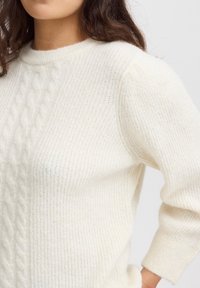 Pull léger en crème avec un motif en maille torsadée texturé. La conception présente un col rond et des manches longues avec des poignets côtelés.