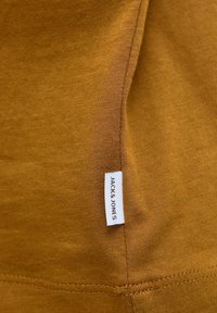 Pamukasto platno boje senfa s glatkom teksturom, s bijelom etiketom s natpisom "JACK & JONES" sašivenom na bočnom šavu.