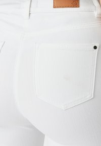 Primer plano de una tela de mezclilla blanca que muestra un bolsillo trasero con una etiqueta de cuero en la cintura y un pequeño detalle de tachuela negra cerca del bolsillo.