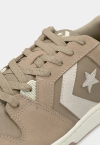 Gros plan d'une basket beige avec des lacets en tissu, une étoile blanche et un logo chevron sur le côté, et une semelle blanche sur fond clair.