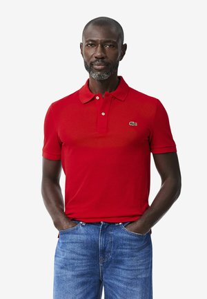 Uomo con barba grigia che indossa una polo rossa con un piccolo logo verde e jeans blu, mani nelle tasche, di fronte.