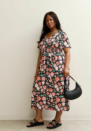 Frau in einem schwarzen, blumigen Midi-Kleid und schwarzen Sandalen, die eine schwarze Leder-Handtasche in Halbmondform hält, steht vor einer schlichten Wand.