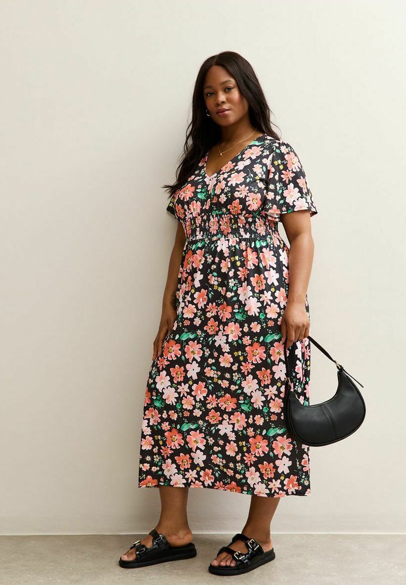 Mujer vestida con un vestido midi negro de flores y sandalias negras, sosteniendo un bolso de cuero negro en forma de media luna, de pie contra una pared lisa.