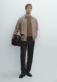 Braune Wollmisch-Jacke mit Reißverschluss, braunes T-Shirt, dunkle Hose, schwarze Schuhe, hält eine schwarze Wildledertasche mit silberner Hardware, steht vor einem hellen Hintergrund.