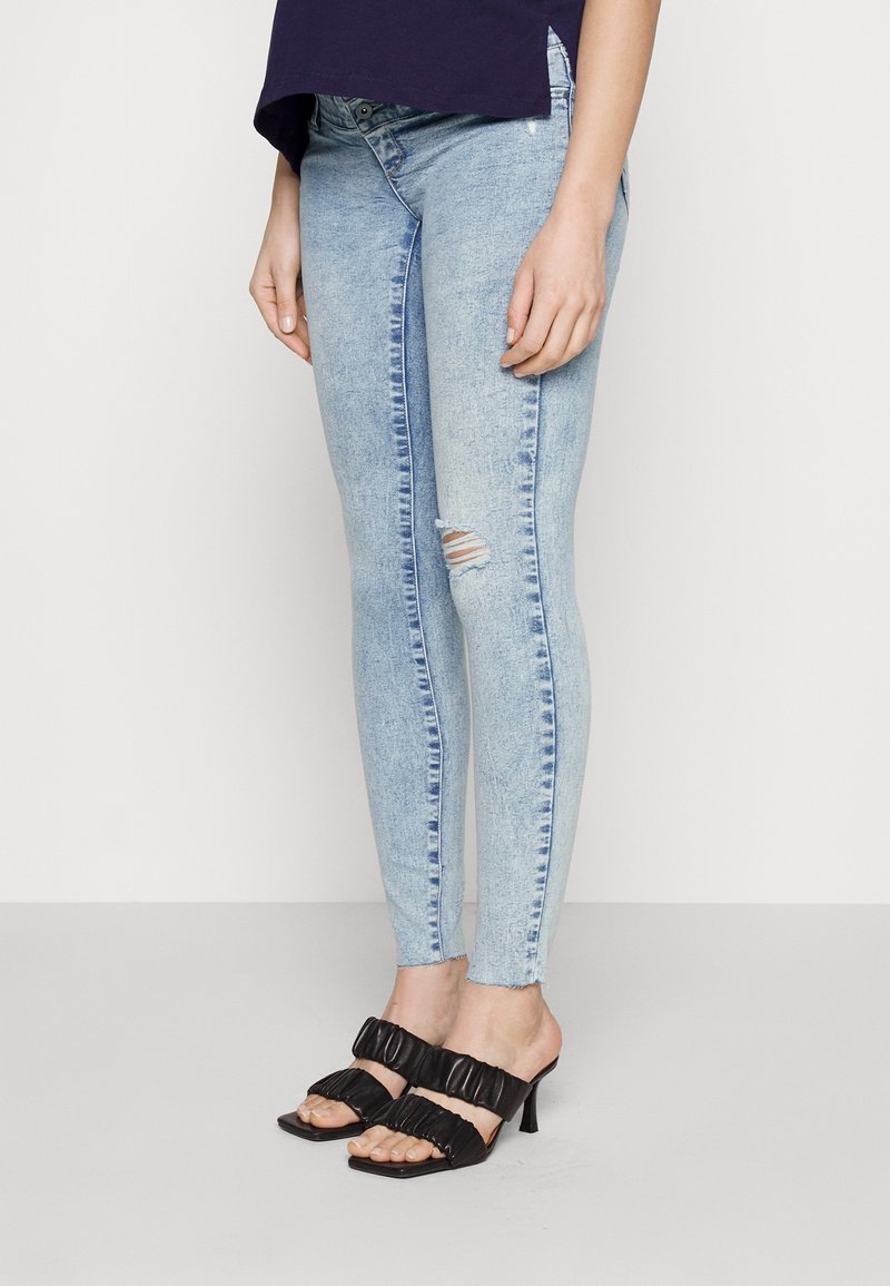 Vaqueros ajustados de mezclilla en azul claro con detalles desgastados, combinados con sandalias de tacón negras con tiras fruncidas y punta cuadrada.