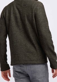 Uomo che indossa un maglione lungo a maniche di un verde scuro e jeans grigi, mostrato di spalle su uno sfondo bianco puro.