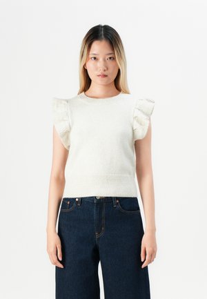 Ungdommelig kvinne står iført en ermeløs hvit strikket topp med volanger på skuldrene og mørkeblå høyhalsede jeans foran en ensfarget bakgrunn.