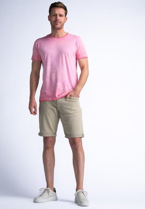 Helles rosa Baumwoll-T-Shirt mit kurzen Ärmeln, kombiniert mit beigen hochgekrempelten Shorts. Abgerundet durch weiße Turnschuhe mit einem schwarzen Akzent.