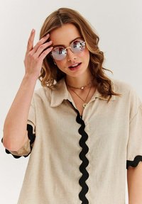 Kulaté sluneční brýle se zrcadlovými čočkami, béžová košile s černým vlnitým lemem, vrstvené náhrdelníky a volné, vlnité vlasy.