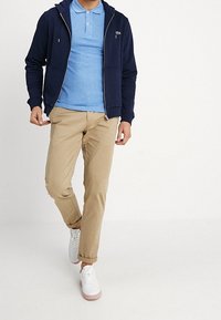 Marinblå zippered hoodie över en ljusblå polo, khakibyxor och vita sneakers med rosa sulor. Har fickor och en liten logotyp på hoodien.