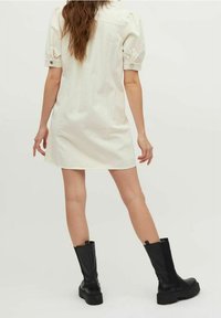 Vaalea beige lyhyt mekko, jossa on puffihihat, nappityylisiä yksityiskohtia ja suora leikkaus, yhdistettynä mustiin polven yli ulottuviin saappaisiin.