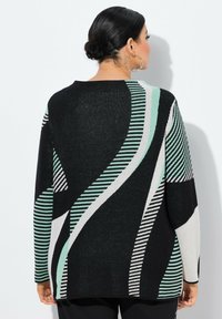 Femme aux cheveux bruns attachés en un chignon bas, portant un pull noir avec des rayures abstraites et courbes en menthe, blanc et noir, se tenant devant un mur blanc.