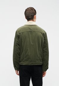 Homme debout de dos, portant une veste en velours côtelé vert olive avec un col en lainage blanc et un pantalon noir sur un fond uni.