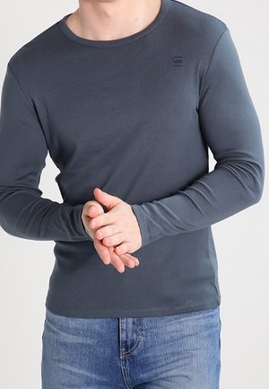 T-shirt à manches longues et col rond en tissu gris foncé, arborant un petit logo sur la poitrine. Texture lisse avec une coupe ajustée. Porté avec un jean bleu.