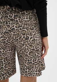 Shorts imprimé léopard avec poches arrière portés par une personne en haut noir à manches longues, montrant le bas du dos et la main droite.