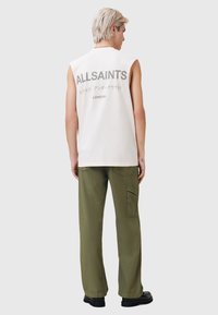 Mouwloos, lichtgekleurde katoenen tanktop met het "ALLSAINTS" logo op de achterkant. Gecombineerd met loszittende olijfgroene broek.