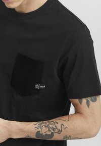 Camiseta negra de manga corta con un bolsillo de parche, que presenta una etiqueta de marca tejida. La tela es suave, con un ligero brillo. Tatuajes visibles en los brazos.