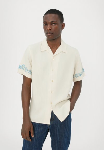 Cămașă cu mânecă scurtă, de culoare crem, cu un guler cu nasturi și broderie florală albastră pe mâneci. Textură fină, croială lejeră, asortată cu blugi.