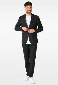 Chaqueta de traje negra con solapas muescas, cierre de un solo botón y acentos azules en los puños. Combinada con pantalones negros ajustados y zapatillas blancas.