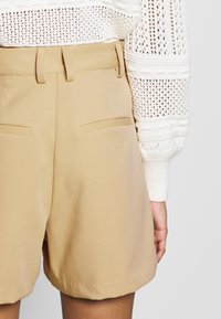 Short beige sur mesure avec passants de ceinture et poches arrière passepoilées, porté avec un pull blanc à manches longues en maille ajourée.