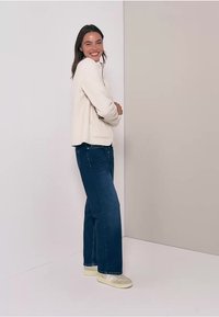 Veste zippée en polaire crème associée à un jean bleu à jambes larges. Le sujet porte des baskets claires et se tient devant un fond neutre.