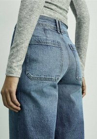 Vaaleansiniset farkut, joissa on rento istuvuus ja näkyvät takataskut. Kankaan pinta on hieman haalistunut ja sileä. Mukana harmaa pitkähihainen paita.