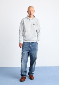 Felpa grigia con cappuccio e logo nero, jeans blu a vestibilità comoda e scarpe in pelle marrone. Tessuto morbido e design semplice.