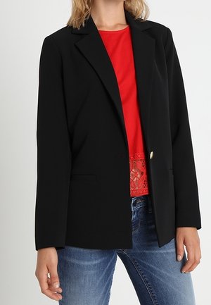 Kvinde iført en sort enkeltknappet blazer, rød top med blonderkant og blå jeans, stående med én hånd afslappet.