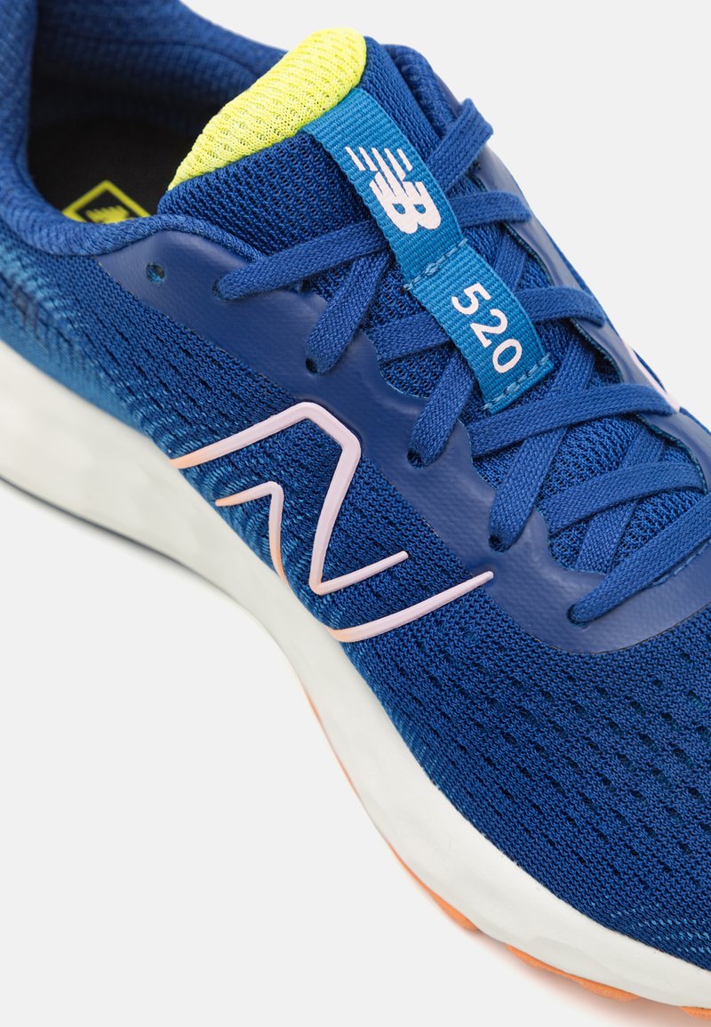 Zapatilla deportiva azul con parte superior de malla, acentos rosas, lengüeta acolchada y suela de goma. Presenta un collar amarillo y una etiqueta de cordón azul con "520".