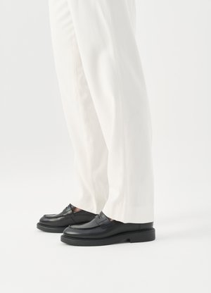 Inimene, kes kannab valgeid rätsepatööna valmistatud pükse ja musti nahast mokasiine, seisab valgel taustal.