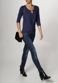 Námořnické tričko s dlouhým rukávem a detaily ve tvaru klíčové dírky, svázané vpředu, v kombinaci s tmavě modrými skinny džíny a černými botami na podpatku.