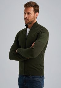 Dunkelgrüner Reißverschluss-Pullover mit hohem Kragen, langen Ärmeln, gerippten Bündchen und einem strukturierten Schulterbereich. Getragen über einem weißen Hemd.