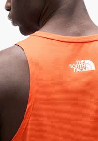 Κοντινό πλάνο ενός ατόμου που φοράει πορτοκαλί αμάνικη μπλούζα με το λογότυπο "The North Face" στο πάνω μέρος της πλάτης.