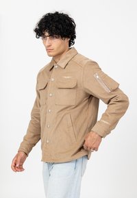 Beżowa kurtka z kordurowego materiału, z zapięciem na guziki, dwiema kieszeniami na piersi i kieszenią na zamek na lewym rękawie. Charakteryzuje się luźnym krojem i teksturowanym materiałem.