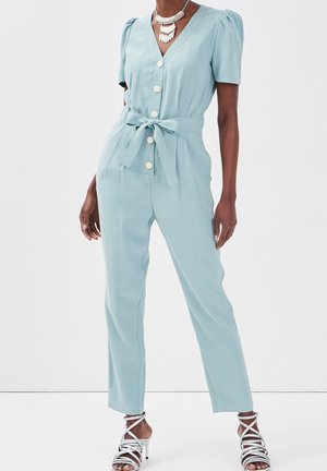 Nainen pukeutuneena vaaleansiniseen haalariin, jossa on lyhyet liehukevät hihat, etunapit, vyötärösolmimisnauha, hopeiset korkeakorkoiset remmisandaalit ja kerroksellinen kaulakoru.