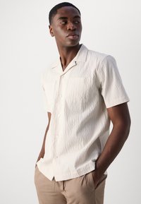 Camisa de manga corta color beige claro con patrón texturizado, diseño con cuello, dos bolsillos frontales y cierre de botones. Lleva puesta con pantalones beige.