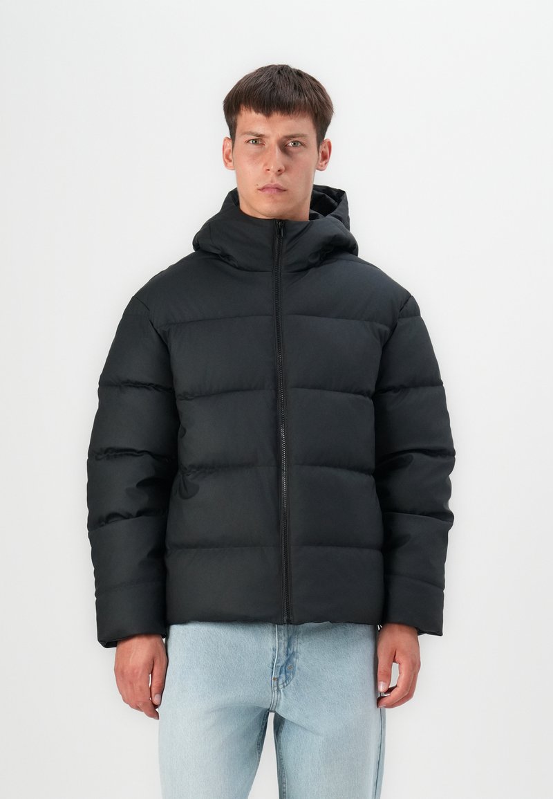 Sort polstret jakke med hætte, med en frontlynlås, glat tekstur og en boxy form. Sammen med lyseblå jeans.