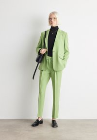 Hellgrüner Anzug mit tailliertem Blazer und Jogginghose mit Kordelzug. Darunter ein schwarzes Rollkragenshirt. Schwarze Schuhe und eine passende Handtasche runden den Look ab.