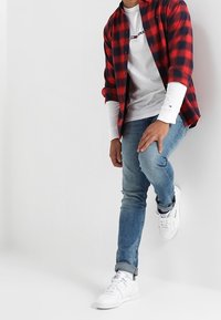Chemise en flanelle à carreaux rouges et noirs sur une chemise à manches longues blanche, associée à un jean bleu et des baskets blanches.