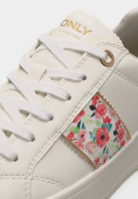 Λευκό αθλητικό παπούτσι με λευκά κορδόνια, χρυσά οπές και λογότυπο, και υφασμάτινο πάνελ με λουλούδια σε κόκκινο, ροζ και πράσινο στη πλευρά.